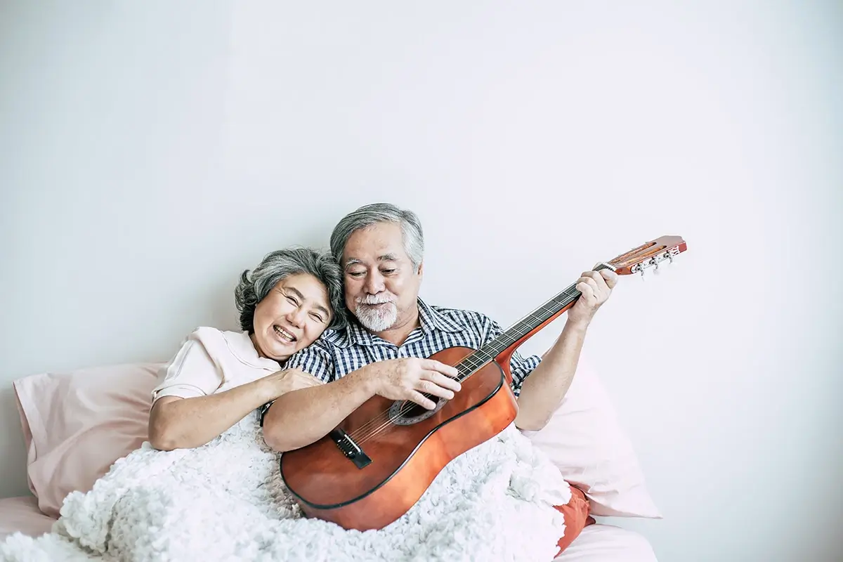An older man playing guitar while his wife hugs his arm